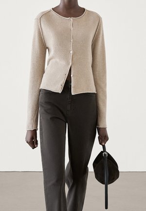 Person iført beige knappet cardigan og mørkegrå bukser, der holder en sort rund håndtaske i remmen med højre hånd.