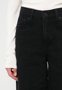 Czarne dżinsy wykonane z teksturowanego denimu, o wysokim stanie, z zapięciem na guzik z przodu, pięcioma kieszeniami i rozsianymi zdobieniami.