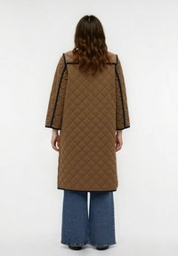 Manteau long matelassé marron avec un motif en losanges, bord bleu et manches larges, associé à un jean bleu évasé. Vue de dos.
