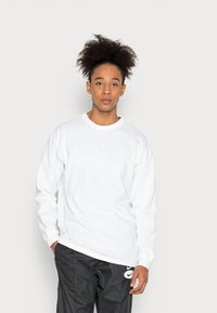 Valge pika varrukaga T-särk, millel on rinnal väike hõbedane Nike logo, koos mustade pükstega. Sile puuvillane kangas, mugav lõige.