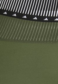 Zelená sportovní látka s texturovanou sítí a pruhovaným vzorem na pasu, s černým lemem a malými bílými logovými akcenty.