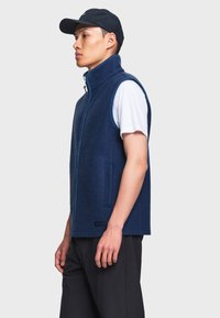 Jeune homme portant une veste sans manches bleu marine zippée, une chemise blanche à manches courtes, une casquette noire et un pantalon noir, debout de profil sur un fond uni.