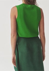 Donna che indossa un top senza maniche in maglia verde brillante e una gonna a vita alta verde scuro, vista da dietro su uno sfondo semplice.