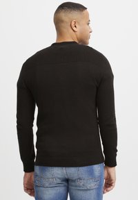 Svart strikkegenser med ribbede detaljer over skuldrene og mansjettene. Har lange ermer og en tettsittende design. Brukt sammen med lyseblå jeans.