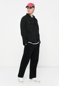 Black corduroy shirt with two flap pockets, worn over a white t-shirt. Black wide-leg pants and white sneakers with beige accents. Red cap with black brim.