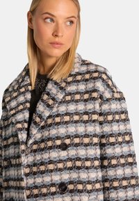 Abrigo de doble botonadura con un patrón texturizado de cuadros en negro, azul claro y beige, con botones en contraste y un cuello entallado.