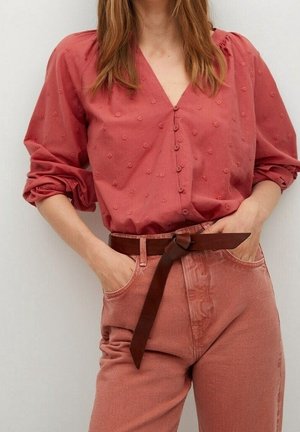 Vrouw draagt een rood gestructureerd blouse met knopen en opgerolde mouwen, roze spijkerbroek en een bruine riem geknoopt, handen in de zakken.