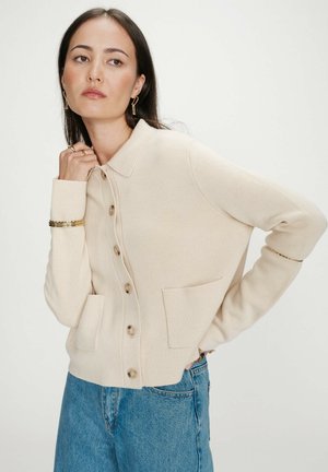 Femme portant un cardigan beige boutonné avec des poches plaquées et un jean bleu, posant avec une main près du cou et l'autre sur la hanche.
