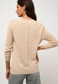 Maglione beige lavorato a maglia con scollo rotondo, maniche a tre quarti e dettagli con bottoni lungo la schiena. Texture a costine sul fondo e sui polsini.