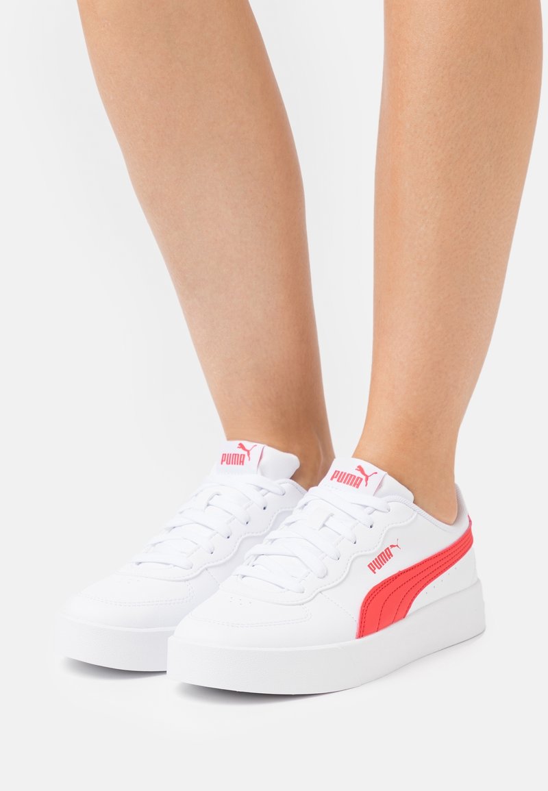Baskets blanches Puma avec bande latérale rouge et logo portées aux pieds sur un fond clair uni.