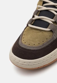 Sneaker con un design multicolore caratterizzato da suede in oliva, marrone e grigio. Lacci bianchi, suola in gomma e dettagli perforati sulla punta.
