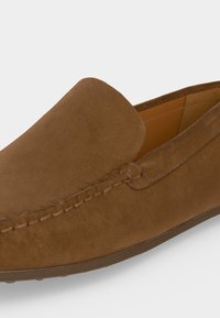 Mocassin en daim marron à enfiler avec des coutures visibles le long du côté et un bout arrondi, vu de côté sur un fond blanc.