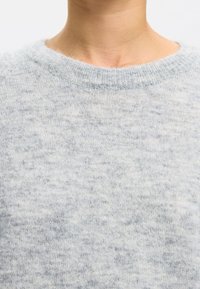 Gros plan sur le cou et la partie supérieure de la poitrine d’une personne portant un pull en tricot doux gris clair avec un col rond.