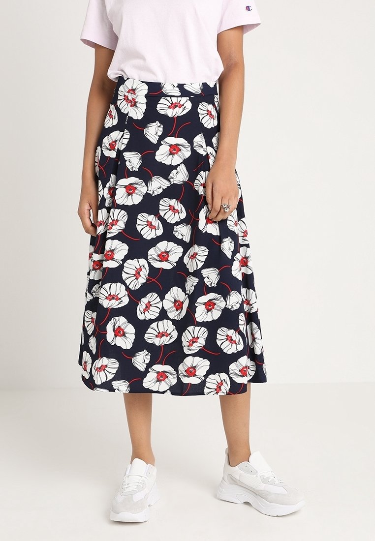 Jupe midi fleurie avec un fond bleu marine, présentant des fleurs blanches aux centres rouges, confectionnée dans un tissu léger. Portée avec des baskets blanches.