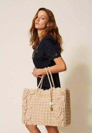 Giovane donna in abito nero che tiene una grande borsa tote beige all'uncinetto con motivi floreali e ciondoli decorativi.