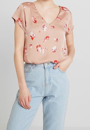 Vrouw draagt een lichtroze bloemen V-hals blouse die in lichtblauwe hoge taille jeans is gestopt, met één hand in de zak.