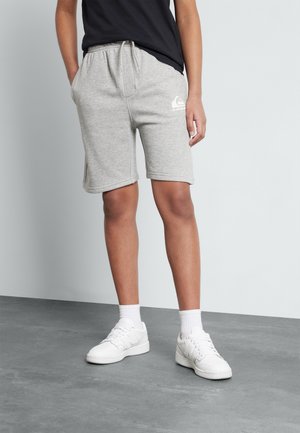 Persona que lleva pantalones cortos deportivos de color gris claro hasta la rodilla con un logo en el muslo izquierdo, calcetines blancos, zapatillas blancas y una camiseta negra.