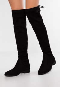 Black suede over-the-knee boots with a fitted design, pointed toe, and low stacked heel. Includes a lace detail at the top.