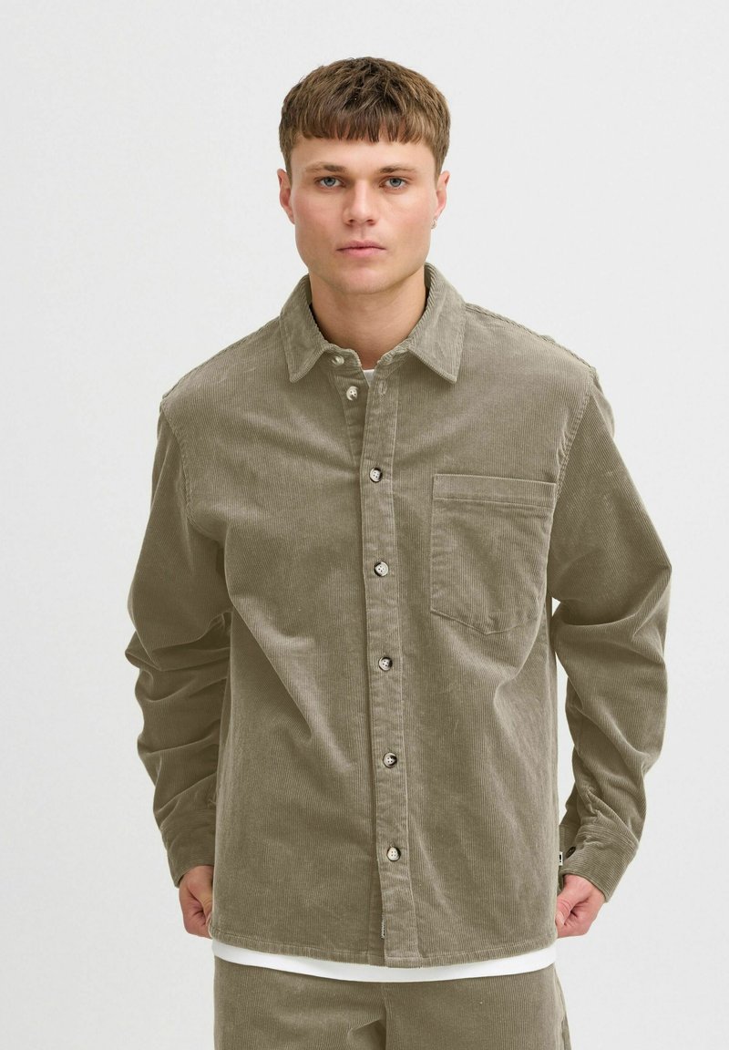 Camicia in costina verde oliva con chiusura a bottoni, colletto e taschino singolo sul petto. Presenta una texture a costine e bottoni argentati.