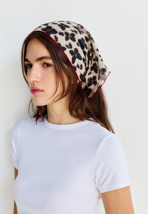 Jeune femme aux longs cheveux bruns portant une chemise blanche et une écharpe beige avec des taches léopard noires et une bordure rouge sur la tête.