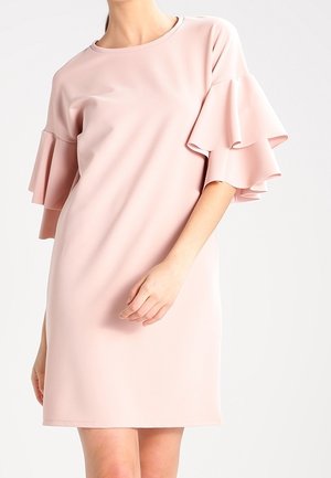 Kobieta w jasnoróżowej sukience do kolan z warstwowymi, falbankowymi krótkimi rękawami na jednolitym tle.