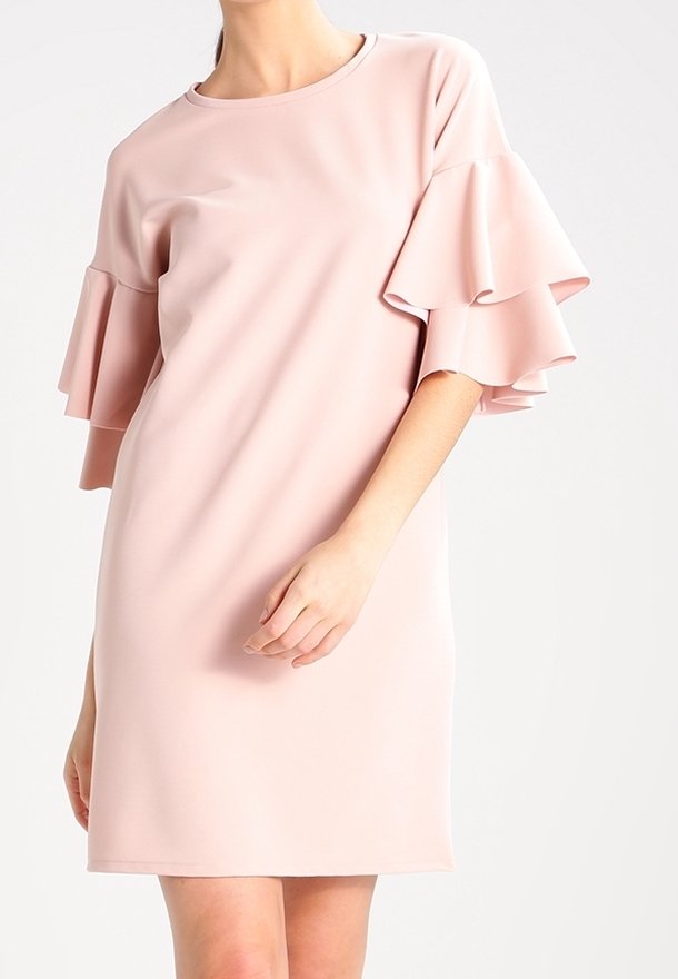 Femme portant une robe rose claire mi-longue avec des manches courtes superposées et volantées, sur un fond uni.