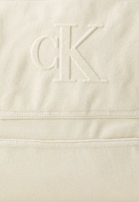 Lerretveske i kremfarge med preget "CK"-logo, to horisontale sømmer og en strukturert stoffoverflate.