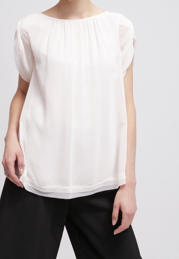 Blusa leggera, trasparente, di colore crema, con scollo rotondo e maniche corte a palloncino, caratterizzata da una leggera arricciatura allo scollo.