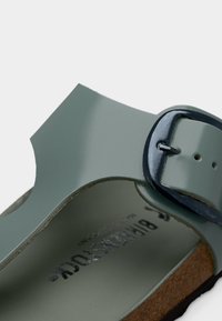 Πράσινο σανδάλι από γυαλιστερό δέρμα με κυρτό, ανοιχτό σχέδιο. Διαθέτει μαύρη αγκράφα και πάτο ποδιού με υφή φελλού. Ομαλές επιφάνειες σε όλο το σανδάλι.