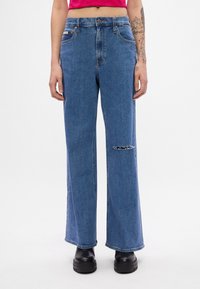 Jeans de ganga largos azuis com uma cintura alta, desbotamento subtil e um detalhe rasgado acima do joelho. Combinados com botas pretas de plataforma.