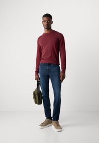 Maroon long-sleeve shirt, blue jeans, and beige sneakers. Model holds a small olive-green bag with a shoulder strap.