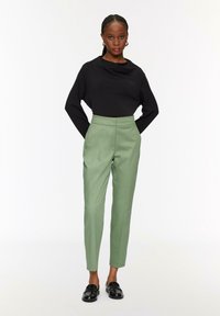 Zwarte gebreide top met een gedrapeerde halslijn, gecombineerd met slim-fit, hooggeplaatste groene broeken. Model draagt zwarte instappers. Effen achtergrond.