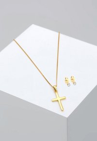 Collana con pendente a croce dorata su una catena, accompagnata da orecchini a bottone dorati con gemme trasparenti. Superficie liscia e riflettente.