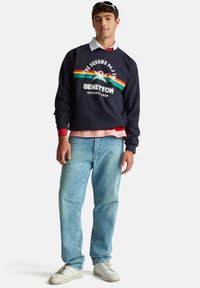 Mladý muž s brýlemi na hlavě, tmavě modrá mikina Benetton s duhovými pruhy, světlé modré džíny, bílé tenisky, stojící na bílém pozadí.