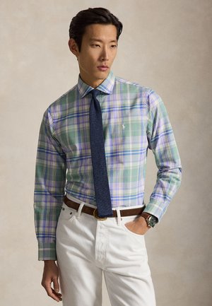 Άνδρας που φοράει πράσινο και μπλε καρό πουκάμισο με σκούρο μπλε γραβάτα, λευκό παντελόνι, καφέ ζώνη και ρολόι, στέκεται μπροστά σε απλό φόντο.