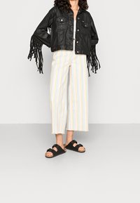Veste cropped en simili cuir noir avec franges, assortie à un pantalon palazzo à rayures blanches et pastel et des sandales à glissière noires.