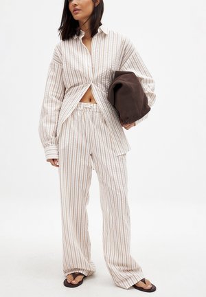 Chemise oversized beige et crème à rayures légères avec un devant boutonné, associée à un pantalon ample assorti et un sac fourre-tout marron.