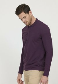 Polo à manches longues de couleur violet profond avec un col texturé, une patte de boutonnage à trois boutons et un logo discret sur la poitrine.