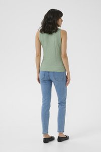 Haut sans manches texturé vert, silhouette ajustée associée à un jean slim bleu clair et des chaussures plates noires. Fond sobre.