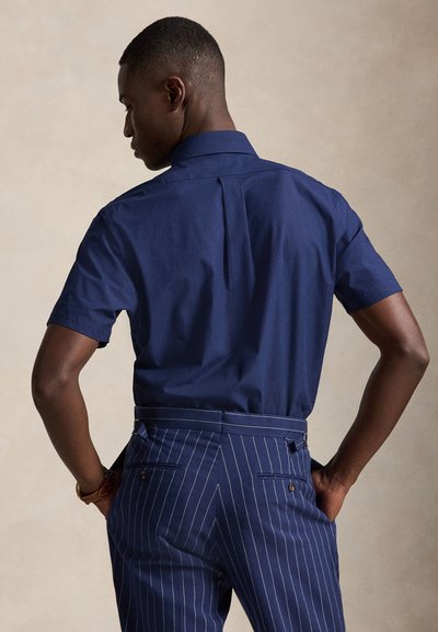 Muž nosiace tmavomodrú krátku košeľu zastrčenú do tmavomodrých nohavíc s tenkými pruhmi, stojí s rukami v bok, otočený chrbtom.