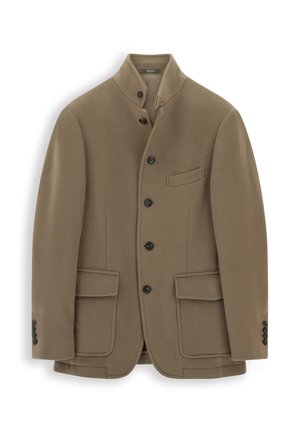 Homme portant des lunettes de soleil, un blazer marron, un pull beige zippé, un pantalon foncé et des gants, tenant un sac à dos en cuir marron.
