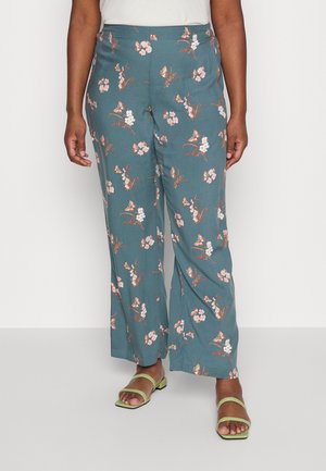 Persoon draagt teal wijde broek met roze en witte bloemenprints en lichtgroene open-toe sandalen, staand tegen een witte achtergrond.