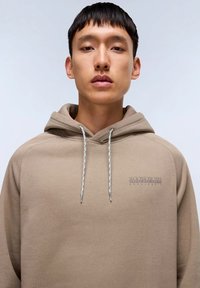 Felpa beige con cappuccio e coulisse, caratterizzata da un tessuto testurizzato e un logo ricamato sul petto. Il design ha una vestibilità rilassata e una tasca a marsupio.