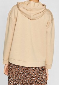 Felpa beige con cappuccio, dal tessuto morbido, con una vestibilità comoda e orlo a costine. Indossata sopra una gonna marrone con motivo leopardato.