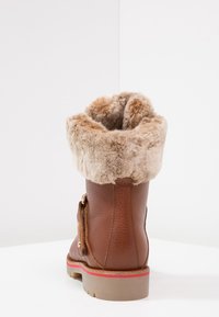 Brun läderstövel med en mjuk, beige fuskpälsupprullning, en sido-spänne och en tan-gummisula med en röd rand.