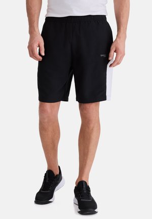 Mann trägt schwarze Sportshorts mit weißen Seitenpanelen und schwarze Laufschuhe, steht vor einem einfarbigen Hintergrund.