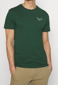 Camiseta de algodón verde de manga corta con un diseño bordado en blanco en el pecho. La tela es suave y tiene un corte relajado.