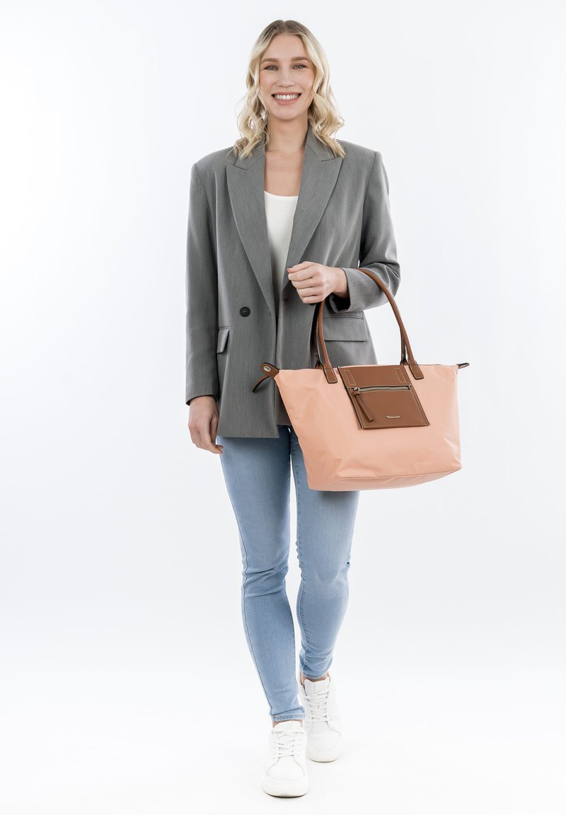 Borsa tote rosa con dettagli in pelle marrone, materiale morbido, design strutturato e una tasca frontale con zip. Il modello indossa un blazer grigio e jeans blu.