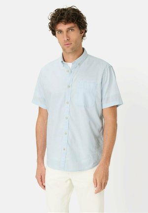 Mann mit lockigem Haar, der ein hellblaues kurzärmeliges Hemd mit Knöpfen und weiße Hose trägt, stehend vor einem schlichten weißen Hintergrund.