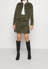 Vrouw in een donkergroene corduroy jas en bijpassende rok, witte coltrui, en zwarte kniehoge laarzen, staande op een grijze vloer.
